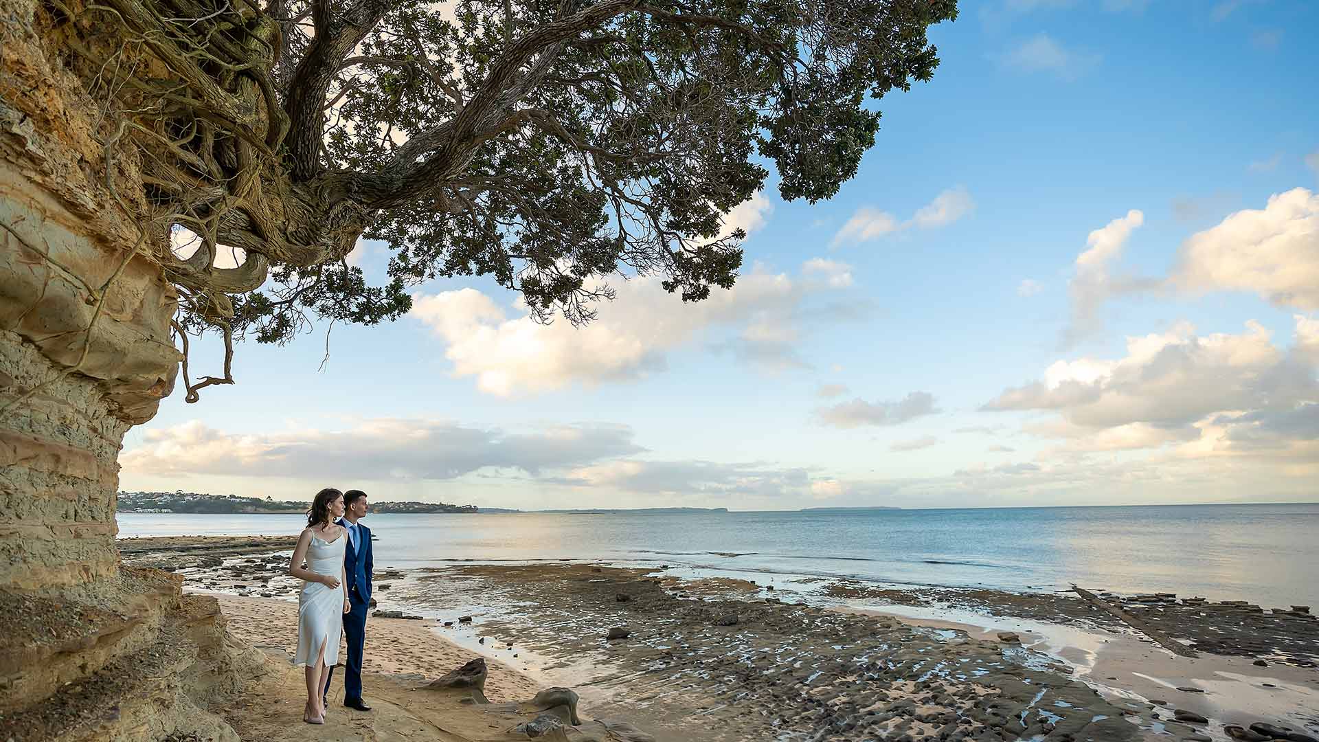 Auckland wedding captured by HAPfilm featuring a couple standing on the beach, with a scenic view of the ocean and sky