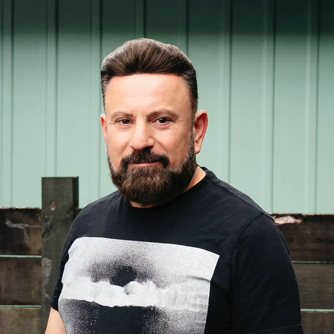 Portrait of Ayad Nuamaan, a professional Assyrian videographer and editor based in Auckland, New Zealand, smiling warmly against a green wooden background.