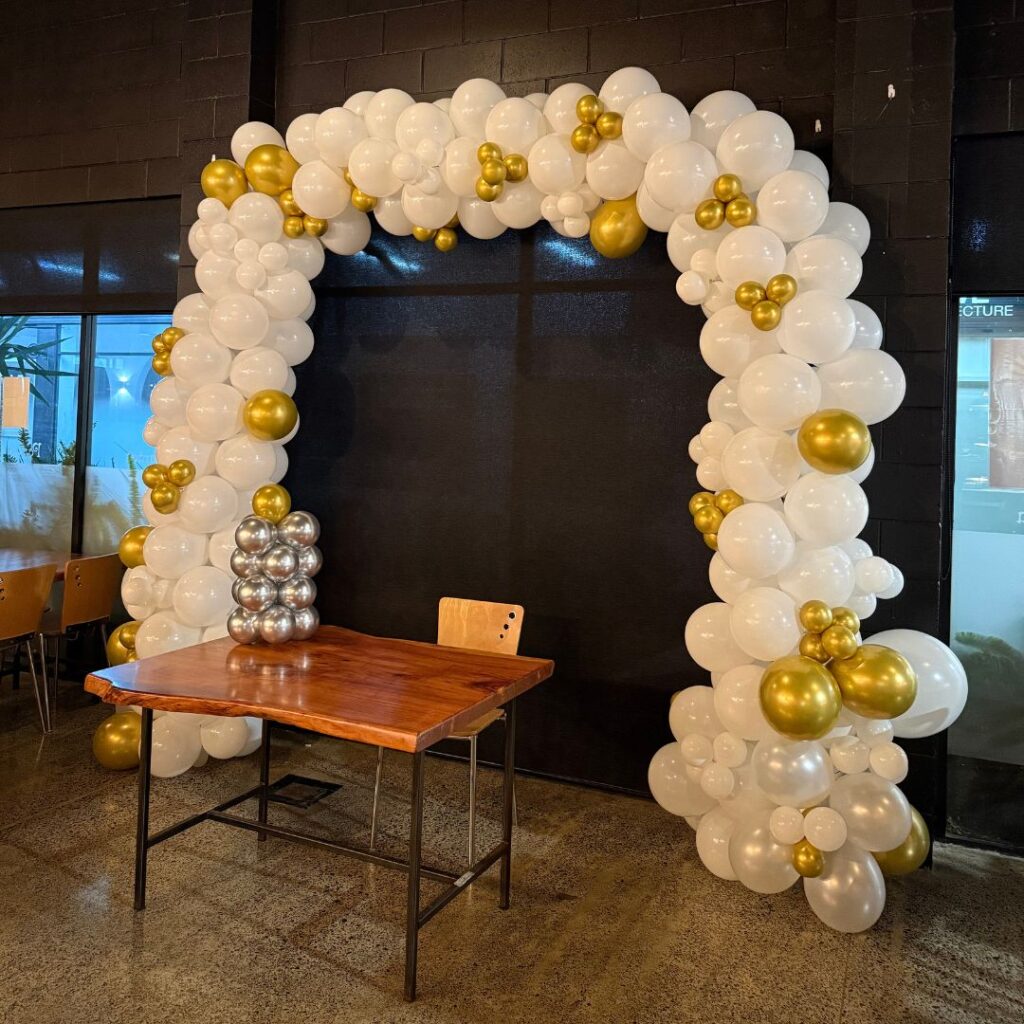 A luxury balloon arch installation in white and metallic gold, set against a black brick backdrop, perfect for an Auckland wedding or corporate event.